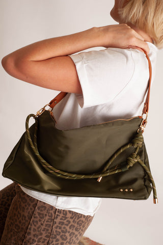 An olive green shoulder bag with a brown leather strap and braided green rope detail, shown being worn over the shoulder of a person in a white t-shirt and leopard print pants.