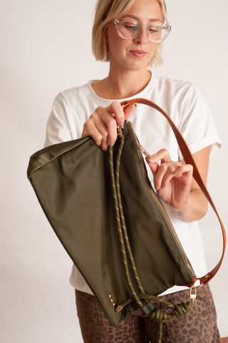 A woman wearing glasses and a white t-shirt holds up an olive green nylon pouch bag with a brown leather strap and drawstring closure against a plain white background.