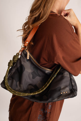 A woman wearing a brown top carries a black camouflage shoulder bag with an olive green braided strap and a tan leather shoulder strap, shown from a side-back angle against a light neutral background.