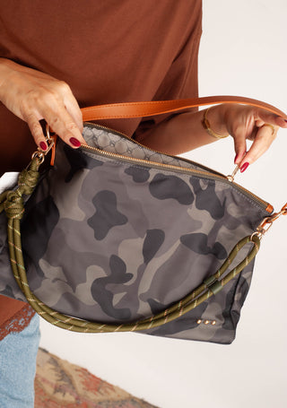 A person holds a gray and black camouflage shoulder bag with a tan leather zipper pull and strap, shown against a plain light background.