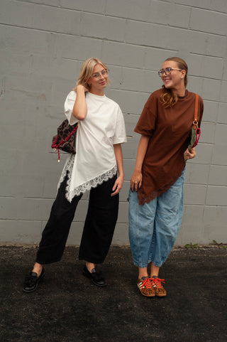 Two women posing outdoors against a gray brick wall, showcasing different casual fashion styles. The woman on the left wears a white oversized t-shirt with a lace-trimmed hem, black wide-leg pants, and black loafers, carrying a small patterned bag.