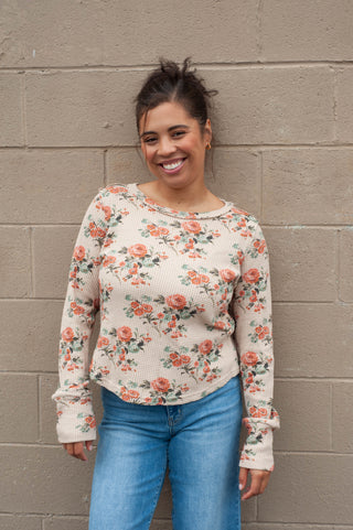 Woman wearing a floral long-sleeve top and blue jeans against a beige brick wall.