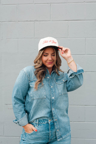 Woman wearing a denim jacket and jeans, holding a white cap with text against a gray wall.