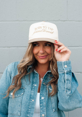 Woman wearing a white cap with text, standing against a gray wall.