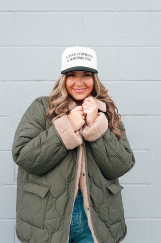 Woman wearing a green quilted jacket and a white cap with text, standing against a gray wall.