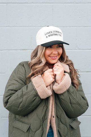 Woman wearing a green puffer jacket and a white cap with text, standing against a gray wall.