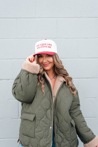 Woman wearing a green quilted jacket and a white cap with text, standing against a light gray brick wall.
