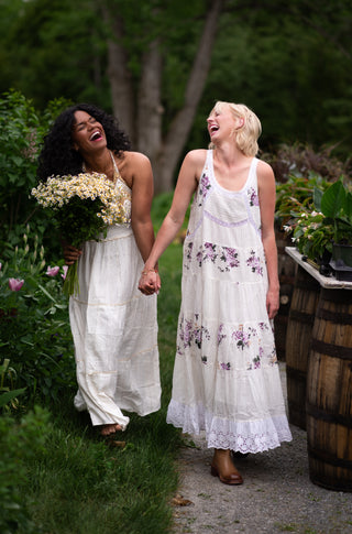 two women wearing white dresses walking and laughing in a garden. 