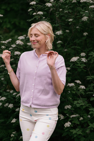 Women wearing a purple cardigan and polka dot pants in front a flower bush.