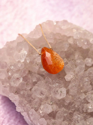 Orange teardrop-shaped pendant on a gold chain against a pink crystal background