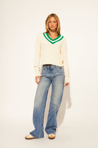 Woman wearing a cream sweater with green trim and blue jeans on a white background