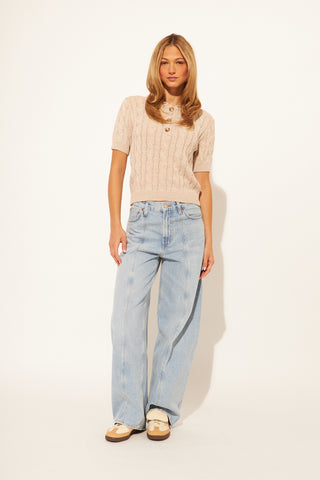 Woman wearing a beige cable knit sweater and light blue jeans on a white background