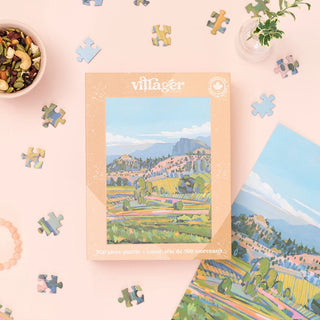 A boxed jigsaw puzzle set by villager, featuring a colorful landscape illustration of vineyard fields and mountains, styled on a light pink background with scattered puzzle pieces and small decorative props.