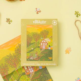 A boxed 500-piece village-themed puzzle from villager, shown standing upright with part of the puzzle and loose puzzle pieces arranged around it on a pale yellow background.