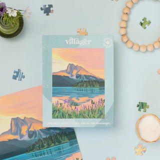 A jigsaw puzzle box labeled 'villager' is centered on a light blue surface, showing a mountain lake landscape with a sunset sky. Additional puzzle pieces, a bead bracelet, a small plant, and a bowl are arranged around it in a styled flat-lay.