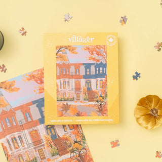 A 1000-piece jigsaw puzzle box by villager, featuring an autumn-themed illustration of colorful Victorian houses, styled on a pale yellow background with scattered puzzle pieces and a small gold pumpkin.