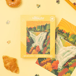A 1000-piece jigsaw puzzle box labeled 'villager' sits on a pale yellow surface, surrounded by puzzle pieces, a croissant, a donut, and part of a pencil. The box features an illustrated waterfall landscape with autumn trees in orange, yellow, green, and brown tones.