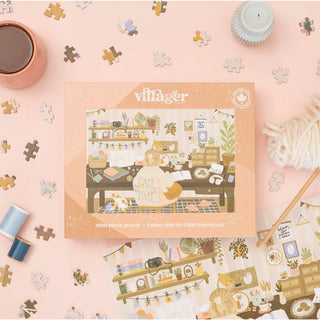 A 1000-piece jigsaw puzzle box by villager, featuring a cozy illustrated shop scene, displayed on a light pink background with scattered puzzle pieces, a cup of coffee, yarn, and other craft-themed props.
