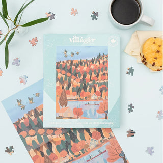 A 1000-piece jigsaw puzzle box titled 'villager' is displayed in a styled flat-lay on a light blue surface, surrounded by scattered puzzle pieces, a cup of black coffee, a biscuit, and green leaves. The puzzle illustration depicts an autumn village scene with colorful trees and ducks.