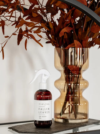 A bottle of Wildflower 'Fallen Leaves' room spray with a white spray nozzle, placed next to a tall, wavy amber glass vase filled with dried brown leaves on a dark slate tray.