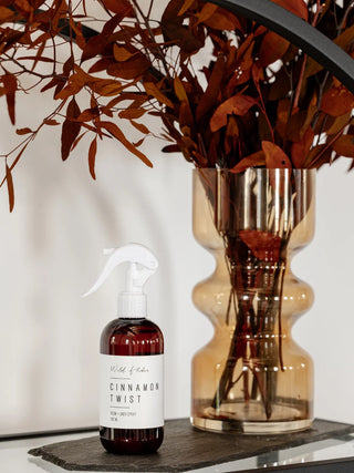 Spray bottle labeled 'Cinnamon Twist' next to a vase with dried leaves on a reflective surface.