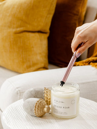 A hand holds a pink lighter over a glass jar candle labeled 'Wildflower Spiced Pear' on a white table, with yellow and brown pillows in the background.