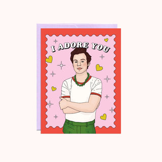 Card with a cartoon man on it with white text saying I Adore You, on a pink background with hearts. 