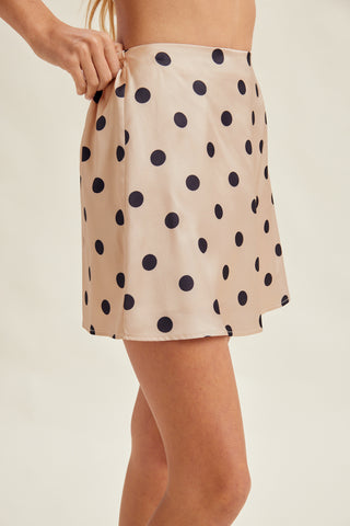A close-up of a model wearing a beige satin-like mini skirt with a black polka dot pattern, holding the waistband with one hand against a plain light beige background.