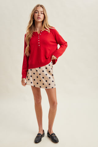 A woman modeling a bright red long-sleeve knit sweater with a buttoned henley neckline, paired with a beige mini skirt featuring black polka dots, and black loafers, against a plain light beige studio background.