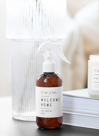 A brown amber spray bottle labeled 'Welcome Home' on a white surface, with a white spray nozzle, surrounded by a white environment suggesting a clean and fresh home atmosphere.