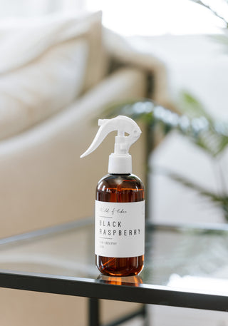 A room linen spray bottle with a white label, containing black raspberry fragrance, placed on a table with a blurred background.