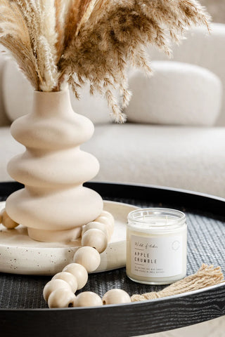 A small glass jar candle labeled 'Wild Bells Apple Scrumble' sits on a black tray beside a beige ceramic vase with dried pampas grass, wooden beads, and a neutral sofa in the background.
