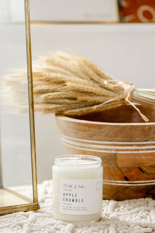 A jar candle labeled 'Wild Flowerz Apple Crumble' sits on a textured cream knit blanket next to a gold-framed mirror and a wooden bowl filled with dried pampas grass.