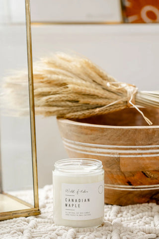 A white soy wax candle in a clear glass jar with a white label reading 'Wildflower' and 'Canadian Maple,' styled on a textured white knit blanket next to a wooden bowl with dried grasses and a gold mirror frame.