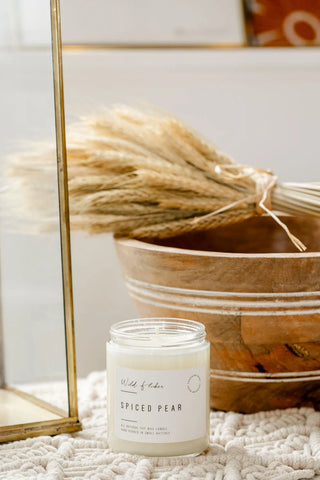 A jar candle labeled 'Wild of London SPICED PEAR' sits on a textured cream knit blanket in a styled home setting, with a wooden bowl, dried pampas grass, and a gold-framed mirror in the background.