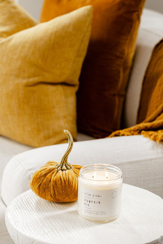 Candle labeled 'Pumpkin Pie' on a small round table with decorative pumpkins and pillows in the background.