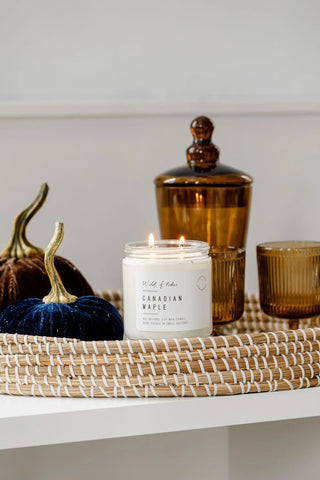 A lit white jar candle labeled 'Wild & Flora Canadian Maple' sits on a woven tray beside amber glass containers and decorative pumpkins on a white shelf.