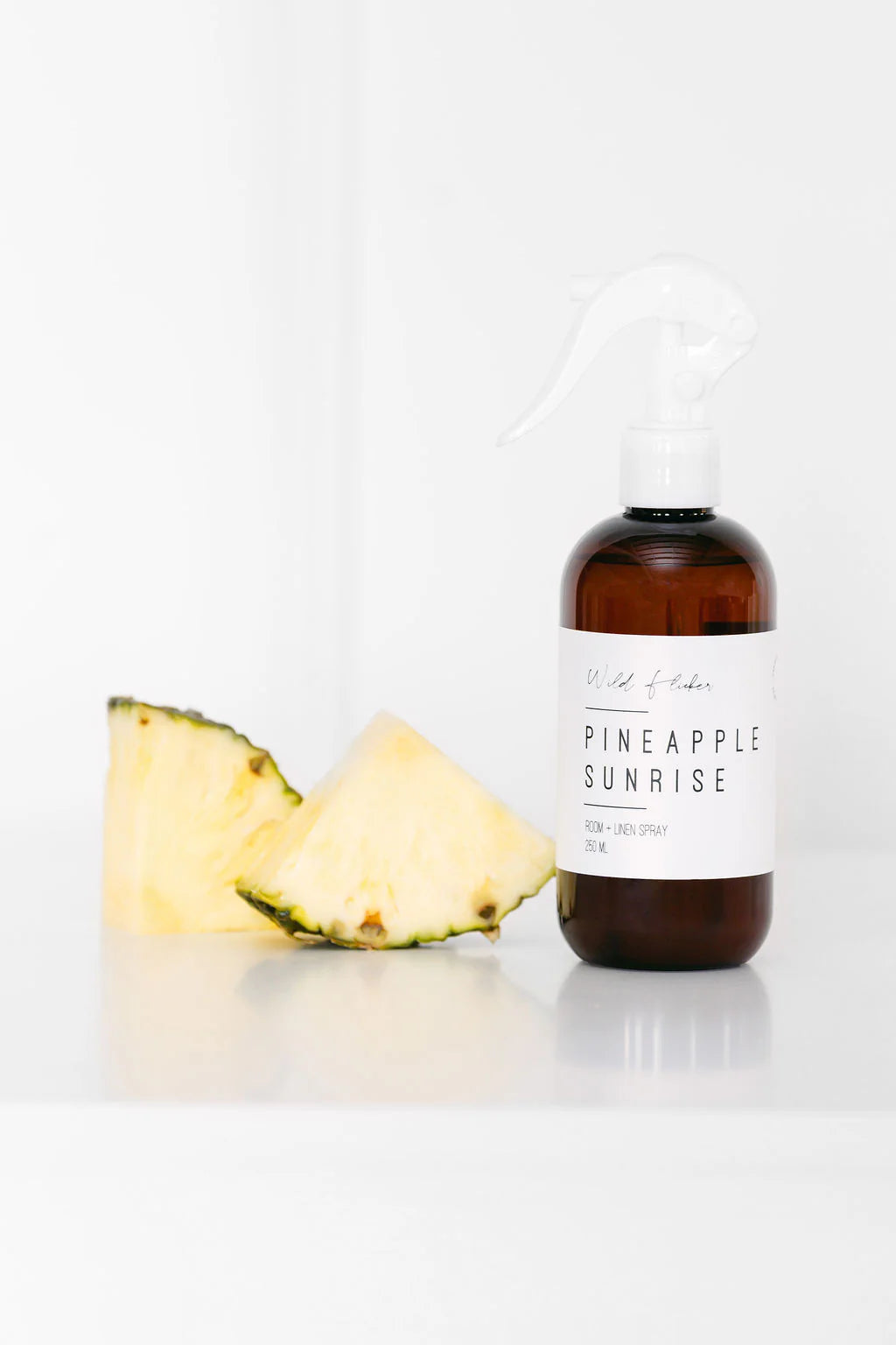 Room & linen spray bottle labeled 'Pineapple Sunrise' next to a pineapple wedge on a white background