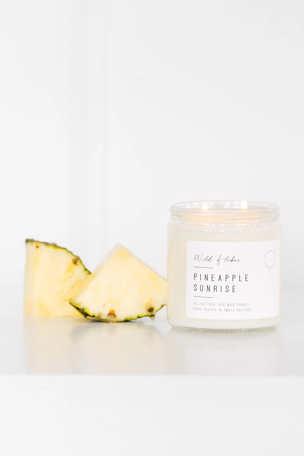 Pineapple candle labeled 'Pineapple Sunrise' next to a pineapple wedge on a white background