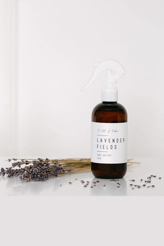 Bottle of 'Lavender Fields' spray on a white surface with lavender flowers.