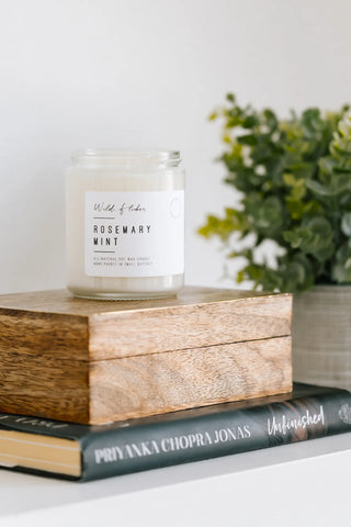 Candle labeled 'Rosemary Mint' on a stack of books with a plant in the background