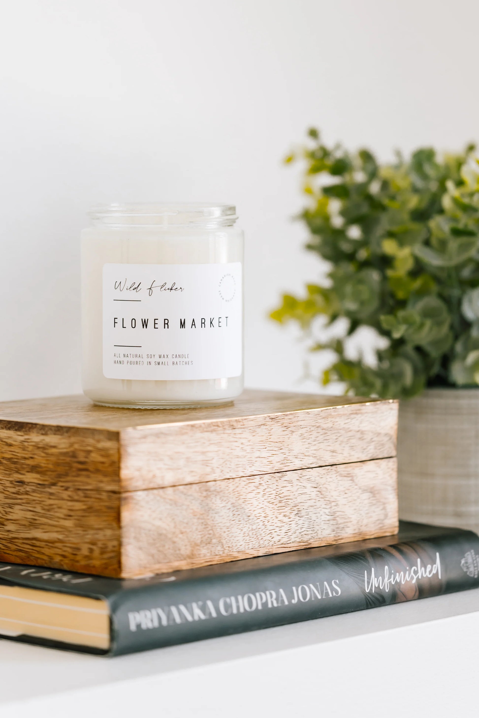 Candle labeled 'Flower Market' on a wooden block with books and a plant in the background