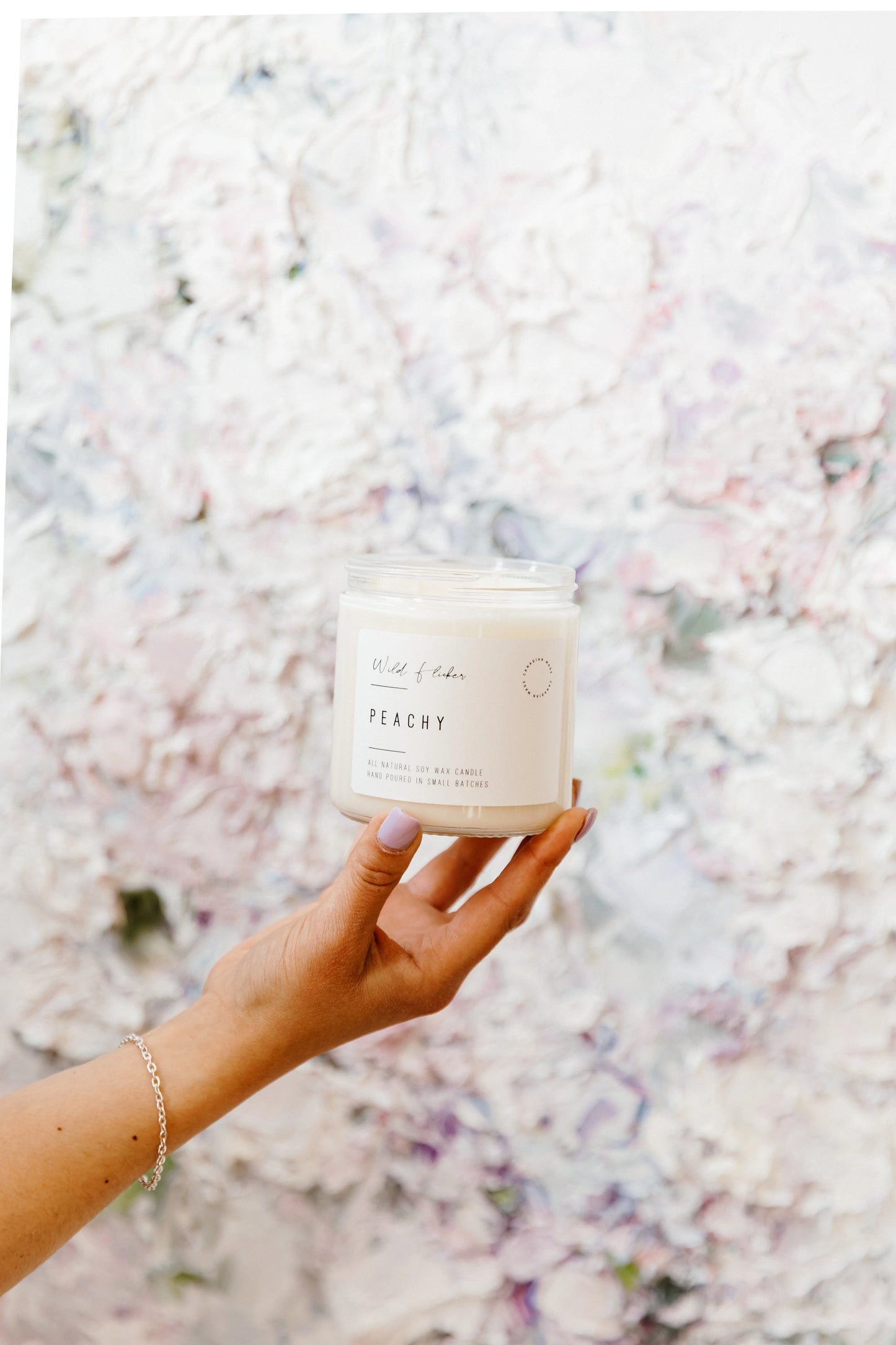 Hand holding a candle labeled 'Peachy' against a floral background