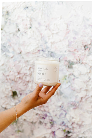 Hand holding a candle labeled 'Peachy' against a floral background