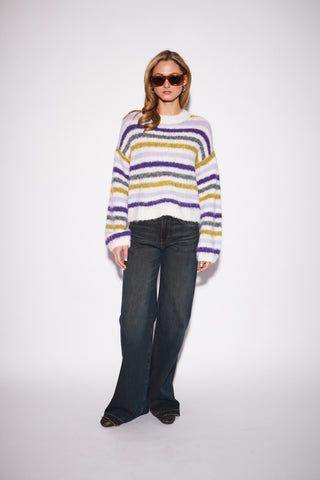 A woman modeling a fuzzy striped knit sweater with dark blue wide-leg jeans and sunglasses, standing against a plain white studio background.