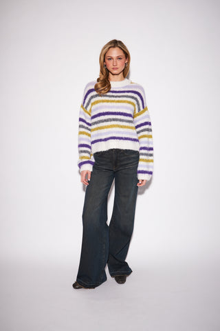 A woman modeling a fuzzy knit sweater with horizontal stripes in white, purple, and mustard yellow, paired with dark blue flared jeans, against a plain light gray studio background.