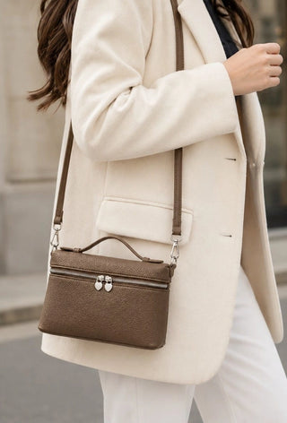 A woman wearing a cream-colored blazer and white pants carries a structured brown leather crossbody handbag with a top handle and silver hardware.