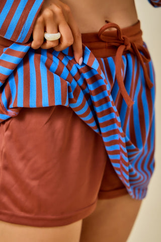Close-up of a person wearing rust-brown shorts and holding a blue and brown striped top or drape over the shorts, with a visible white ring on one finger.