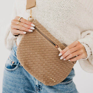 A close-up of a person wearing a beige woven crossbody bag with a gold zipper, styled with a cream knit sweater and blue jeans.