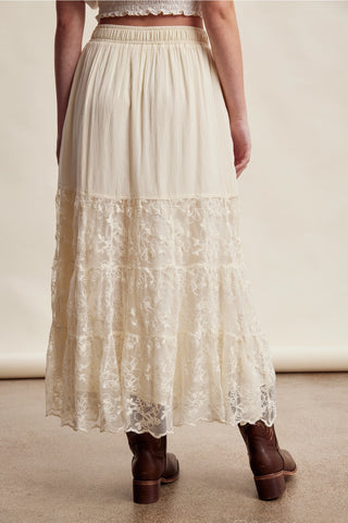 A cream-colored maxi skirt with a lace tiered hem, shown on a model from the back wearing brown leather boots against a neutral beige studio background.